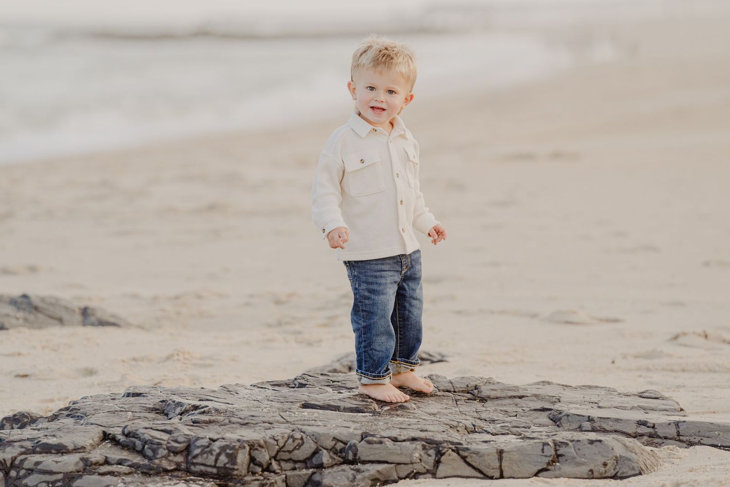 Toddler in white shirt and jeans standing barefoot on rocks at the beach.