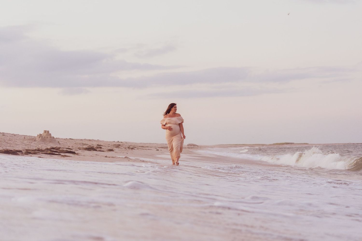 Woman in flowing dress walking along beach shoreline at dusk.
