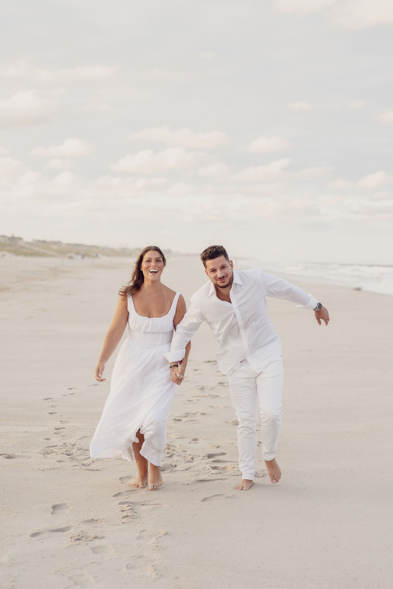 A couple wearing matching white outfits chases each other on a serene beach at sunset.