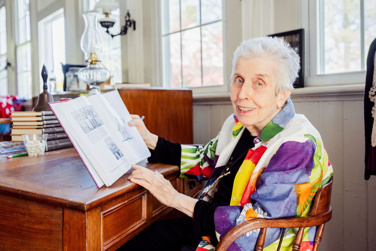 A person in a colorful patterned shirt sits at a wooden desk looking at papers in a sunlit room.