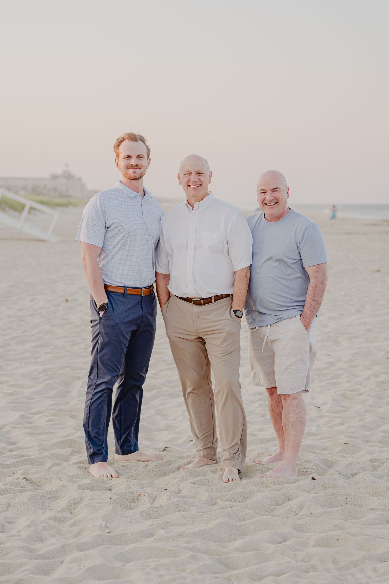 Three smiling men pose together barefoot on a sandy beach at sunset wearing casual light-colored shirts.
