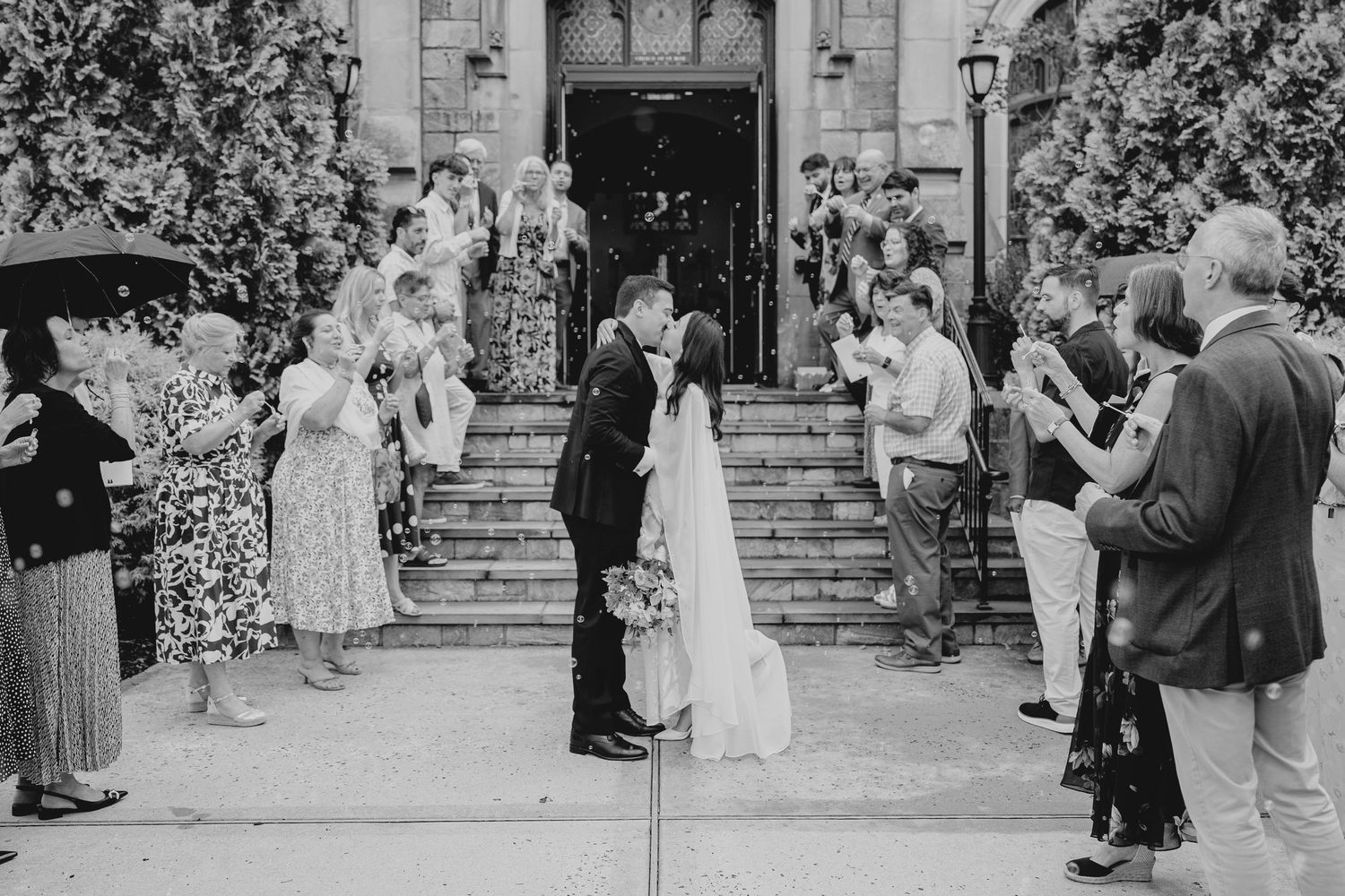 Black and white photo of newlyweds sharing a kiss on church steps surrounded by applauding wedding guests.