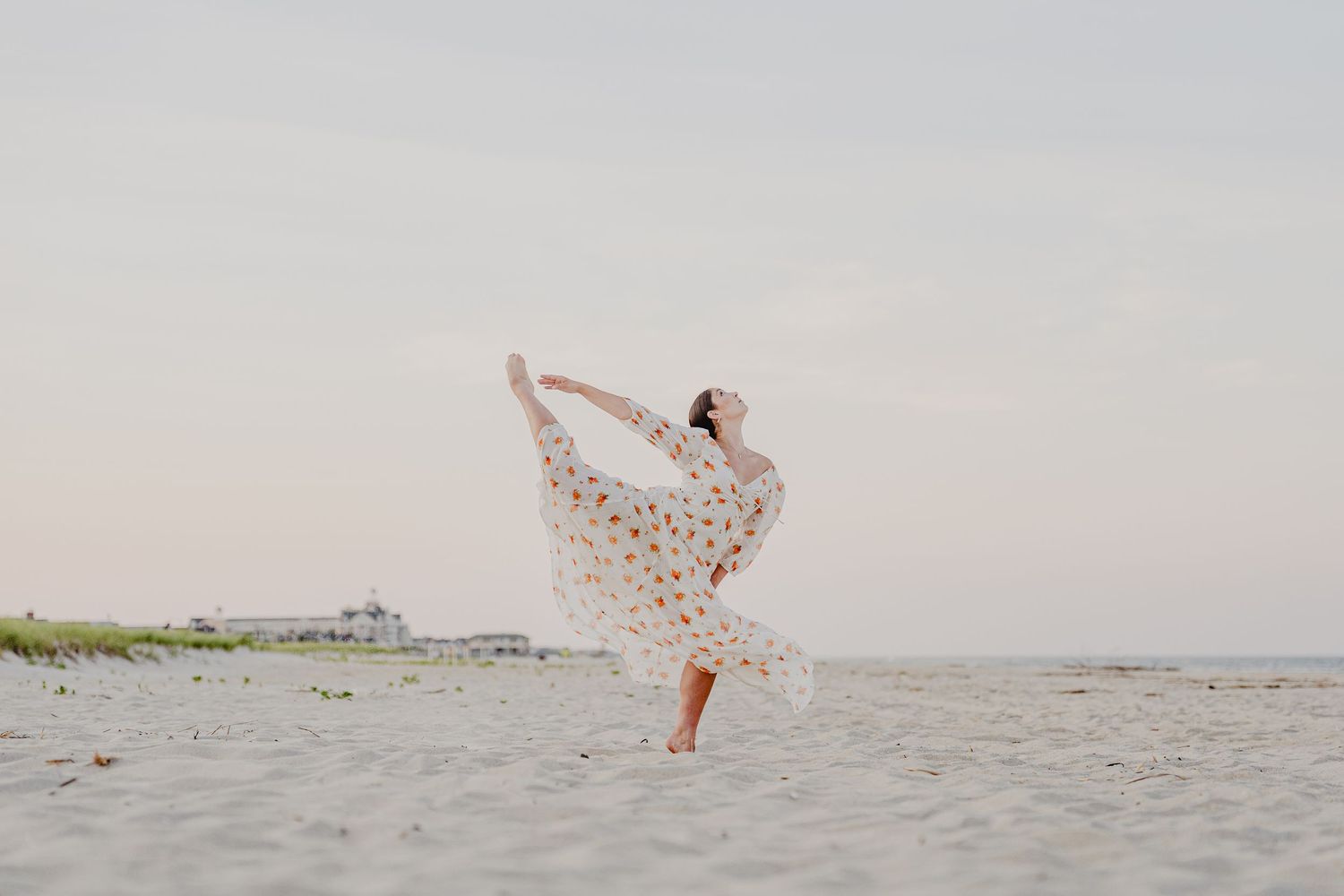 Dancer in flowy polka dot dress performs elegant arabesque on a serene beach at dusk.
