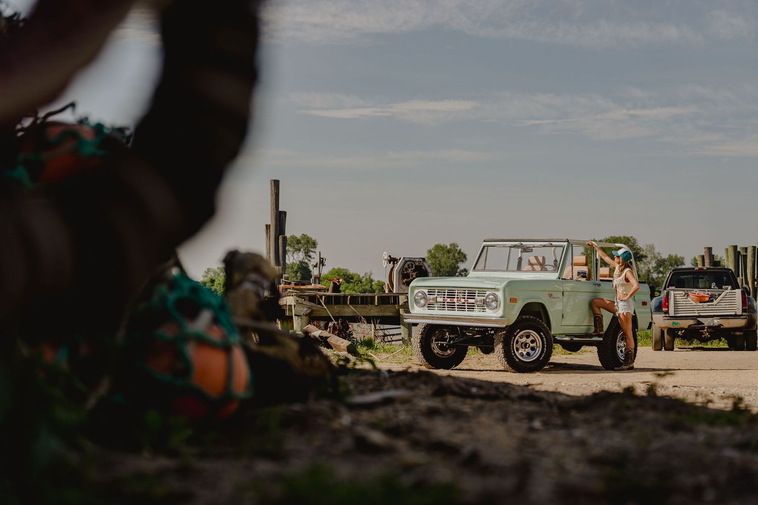 Classic mint Ford Bronco truck parked in muddy field with woman standing beside it on a sunny day.