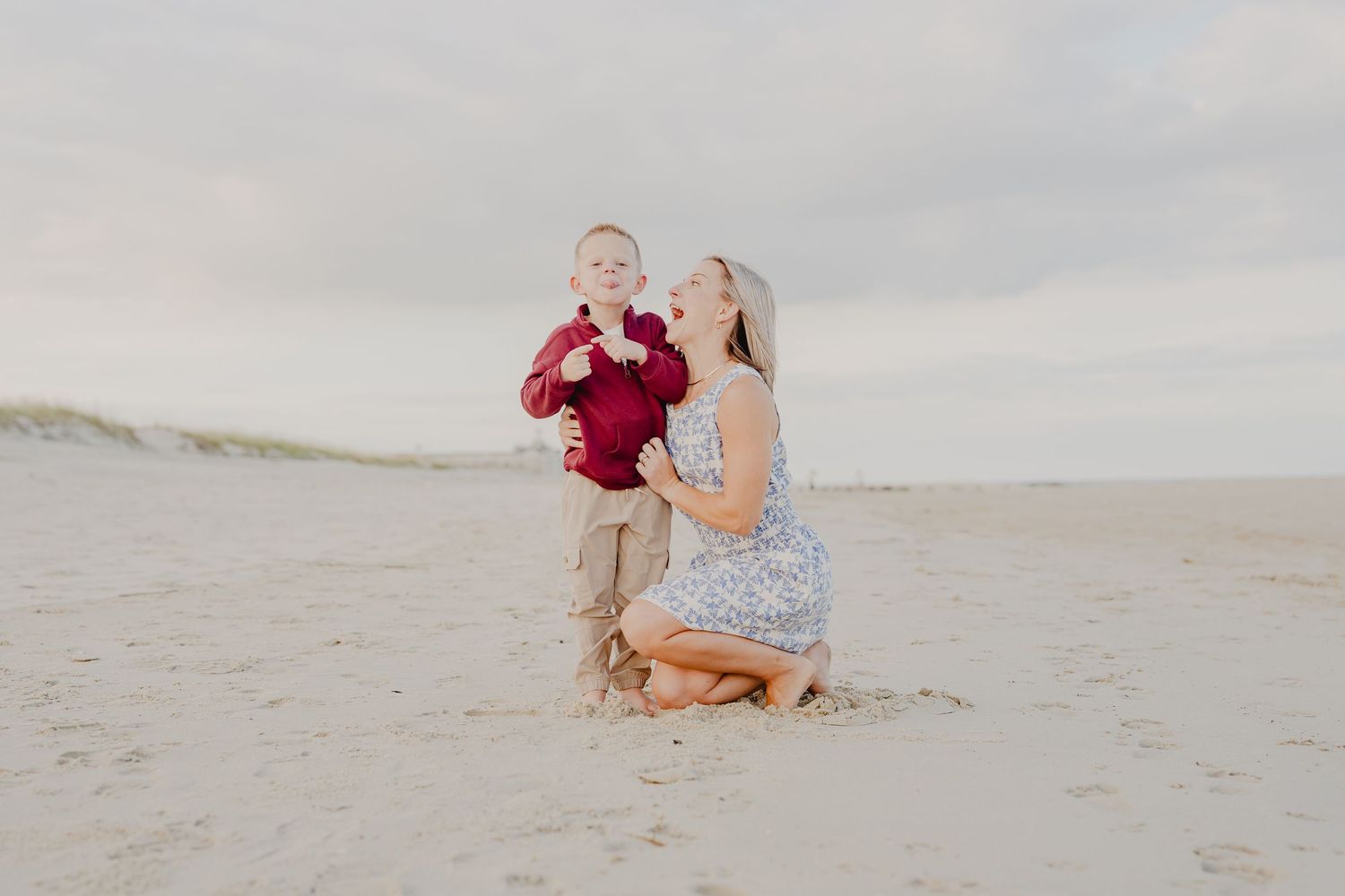 A mother kneels on sandy beach while her young son in red shirt stands beside her, both looking up joyfully.