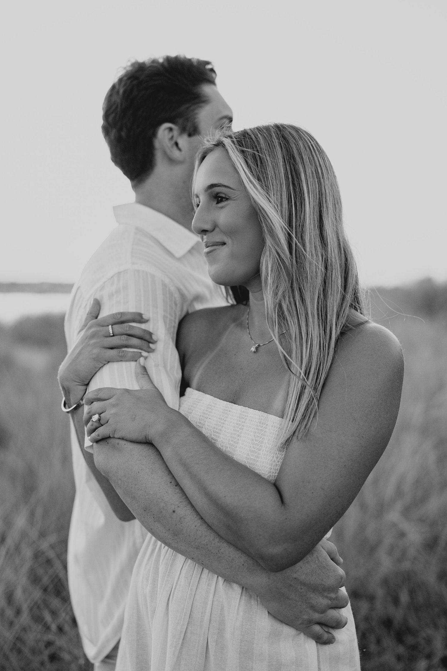 Black and white photo of a couple embracing romantically in a field near water at sunset.