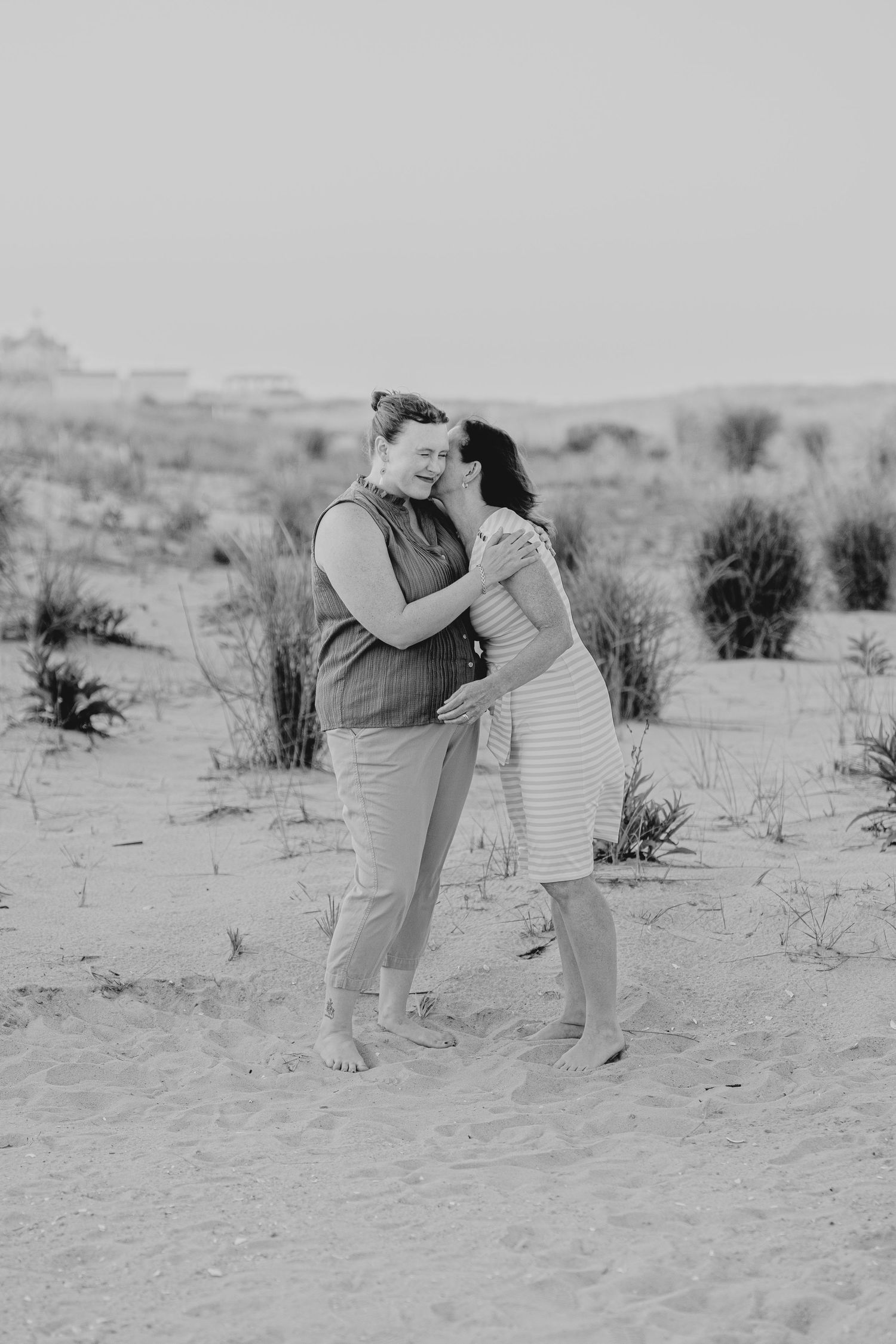 Black and white photo of two women embracing on a sandy beach with dunes and sparse vegetation in the background.