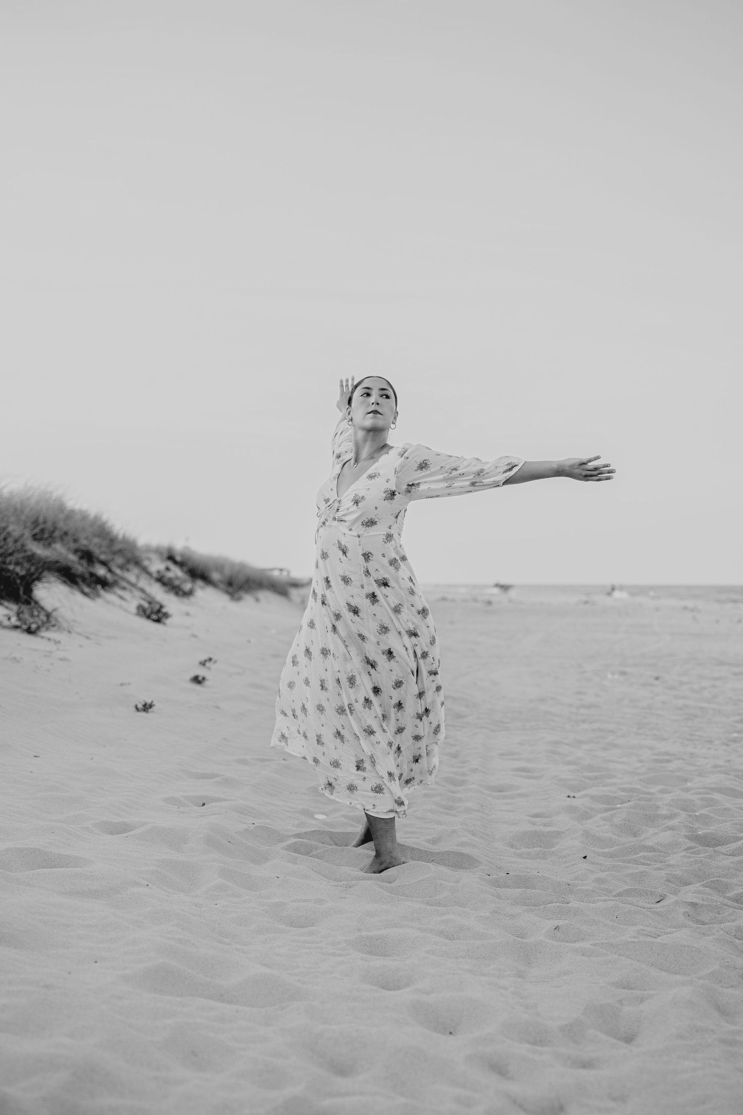 Black and white photo of woman in floral dress with arms outstretched on sandy beach.