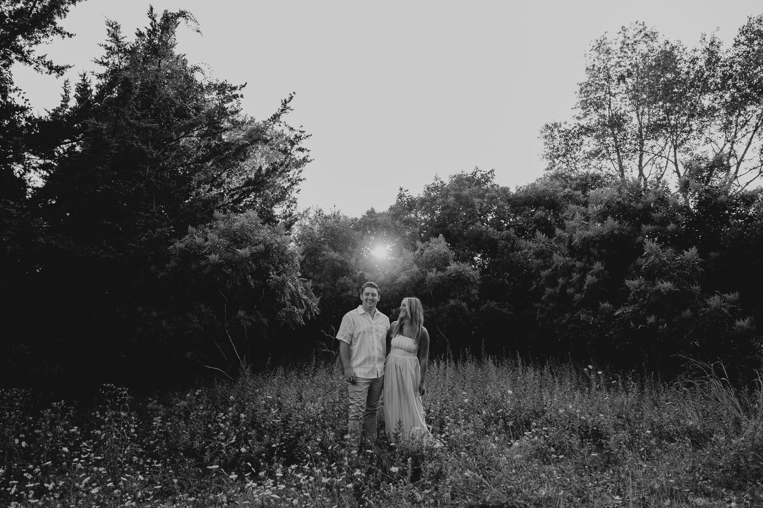 Black and white photo of a couple standing in a wildflower meadow surrounded by trees at sunset.