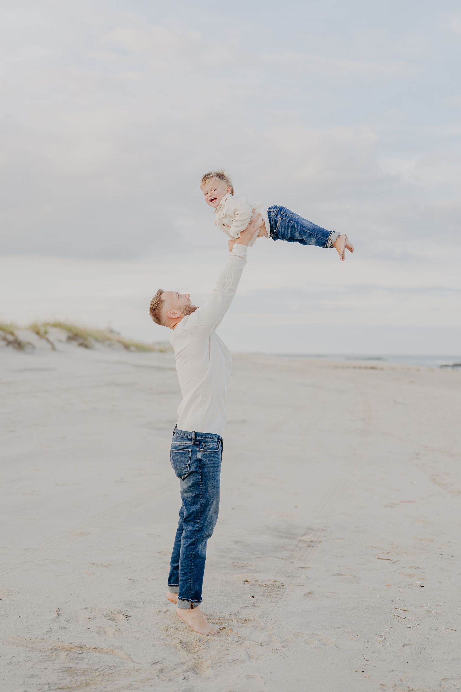 Father joyfully lifts laughing toddler high above his head on a wide sandy beach under cloudy sky.
