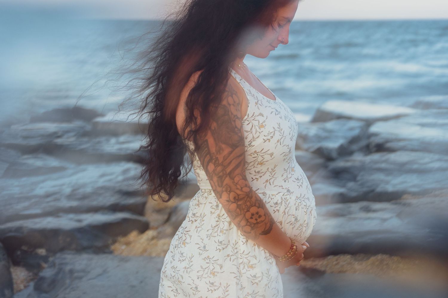 Side view of a pregnant woman in a white dress standing against ocean waves.