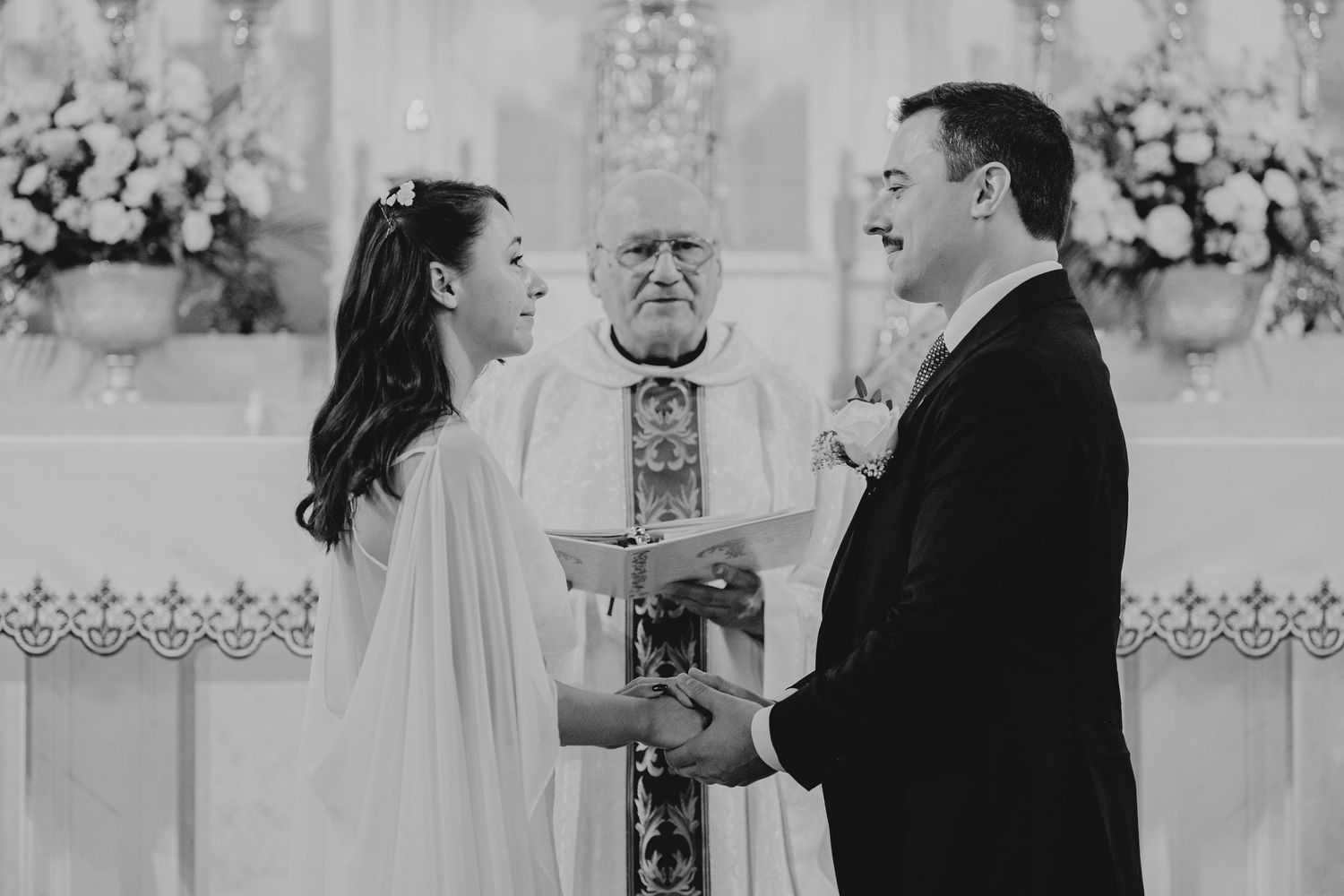 Black and white photo of a bride and groom exchanging vows at a church altar with an officiant behind them.