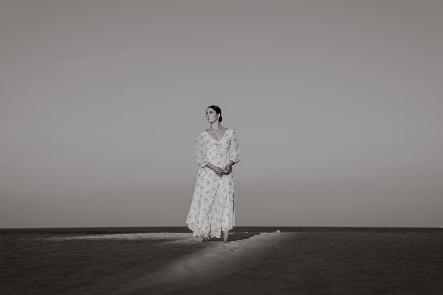 A woman in a flowing white floral gown stands alone on a vast desert plain in a serene black and white photo.