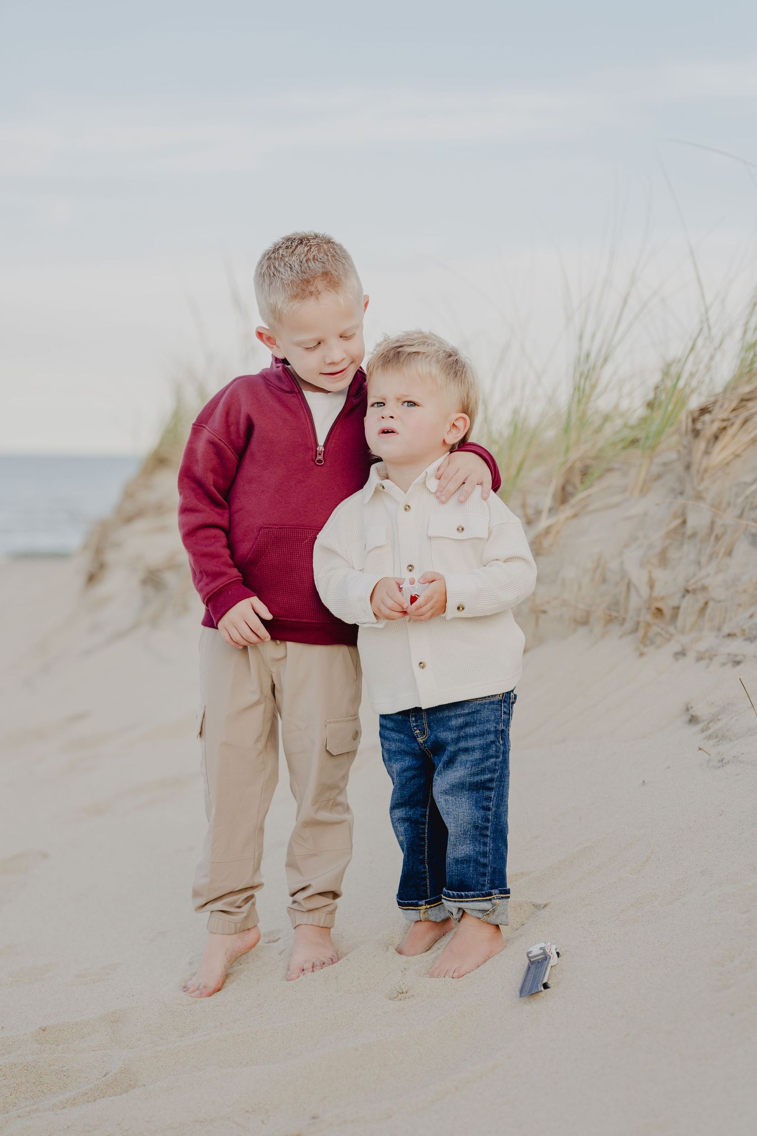 Two young brothers stand barefoot on sandy beach dunes, older boy hugging younger toddler, both smiling outdoors.