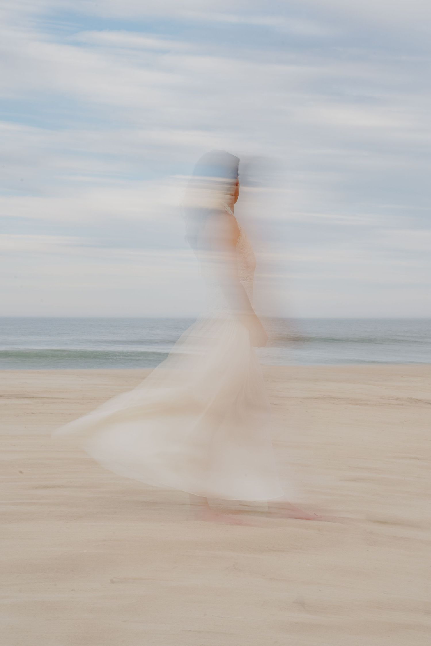 Blurred motion photo of a woman in a white dress walking on a sandy beach with ocean waves in the background.