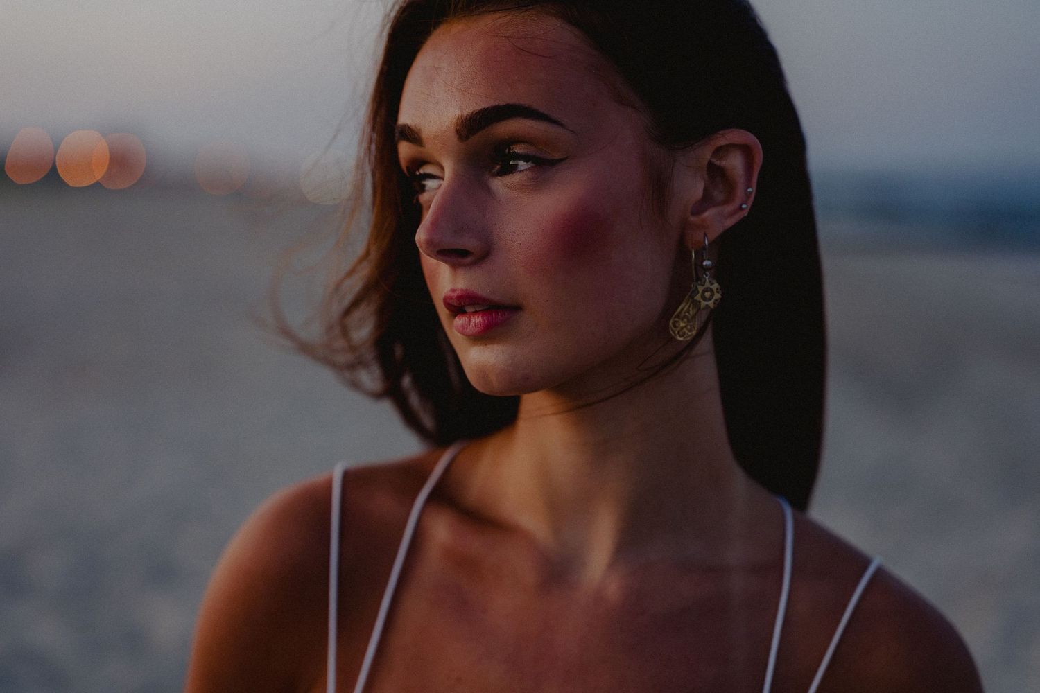 Close-up portrait of young woman with gold earrings and rosy cheeks at beach dusk.