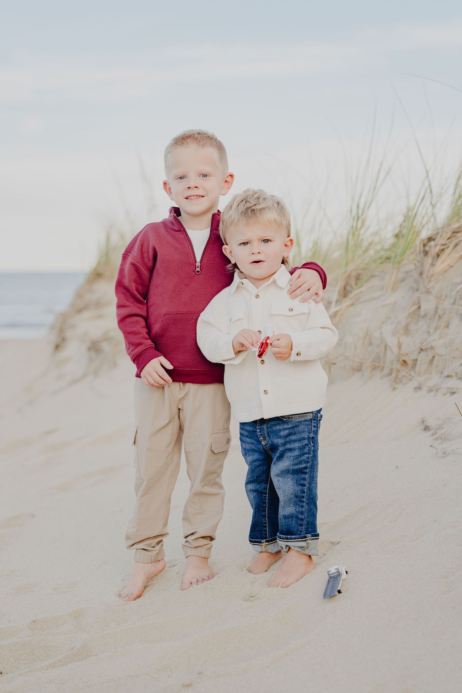 Two young barefoot brothers pose together on sandy beach, older boy in red sweater hugging younger sibling in white jacket.