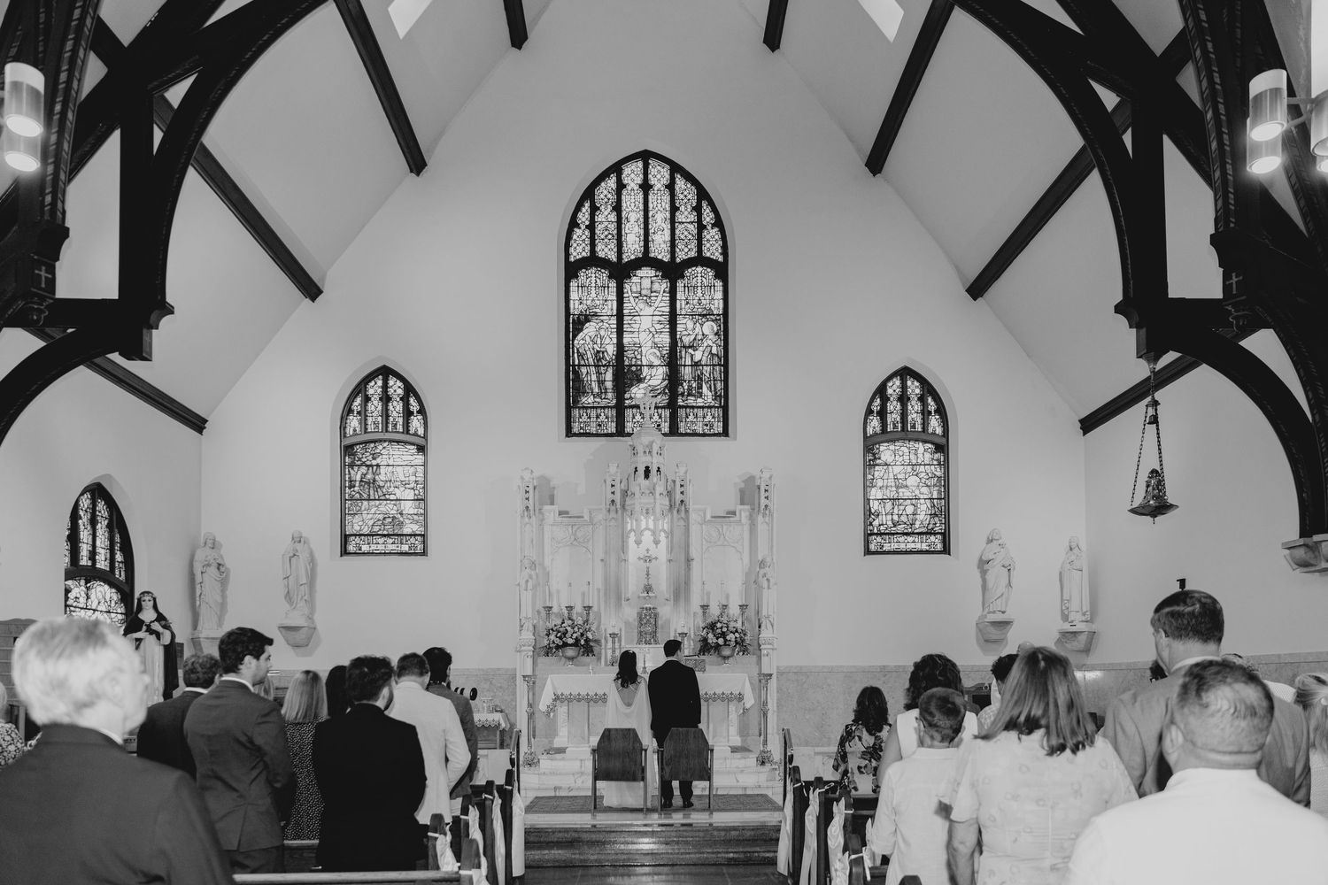 Black and white photo of a wedding ceremony inside a historic church with stained glass windows and wooden pews.
