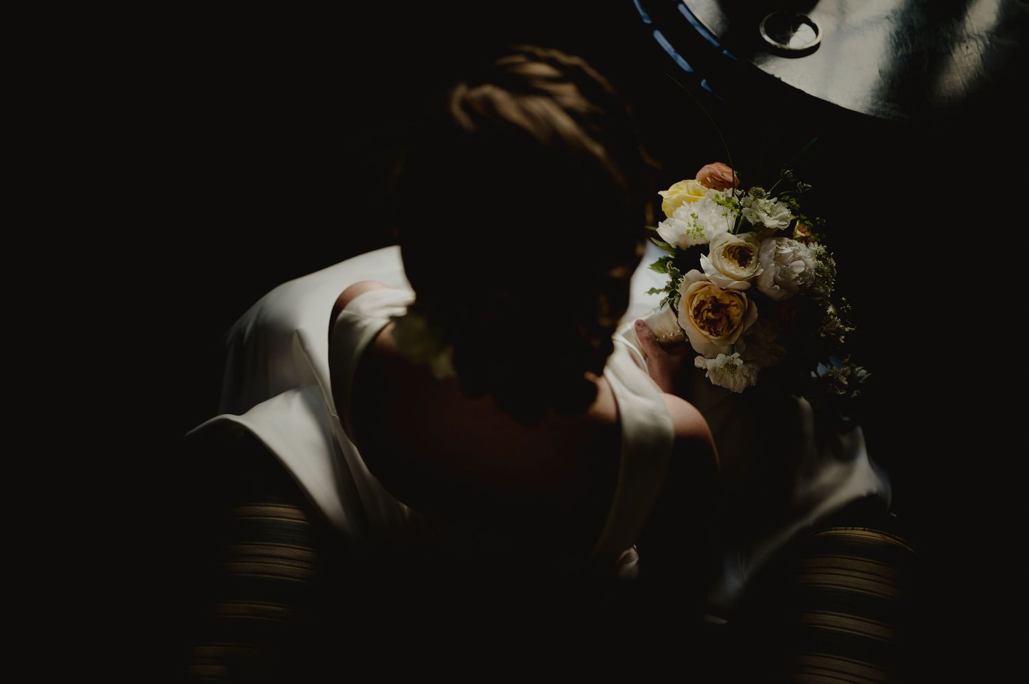 Bride in white dress holding a delicate bouquet of white and peach roses in dramatic dark moody lighting.