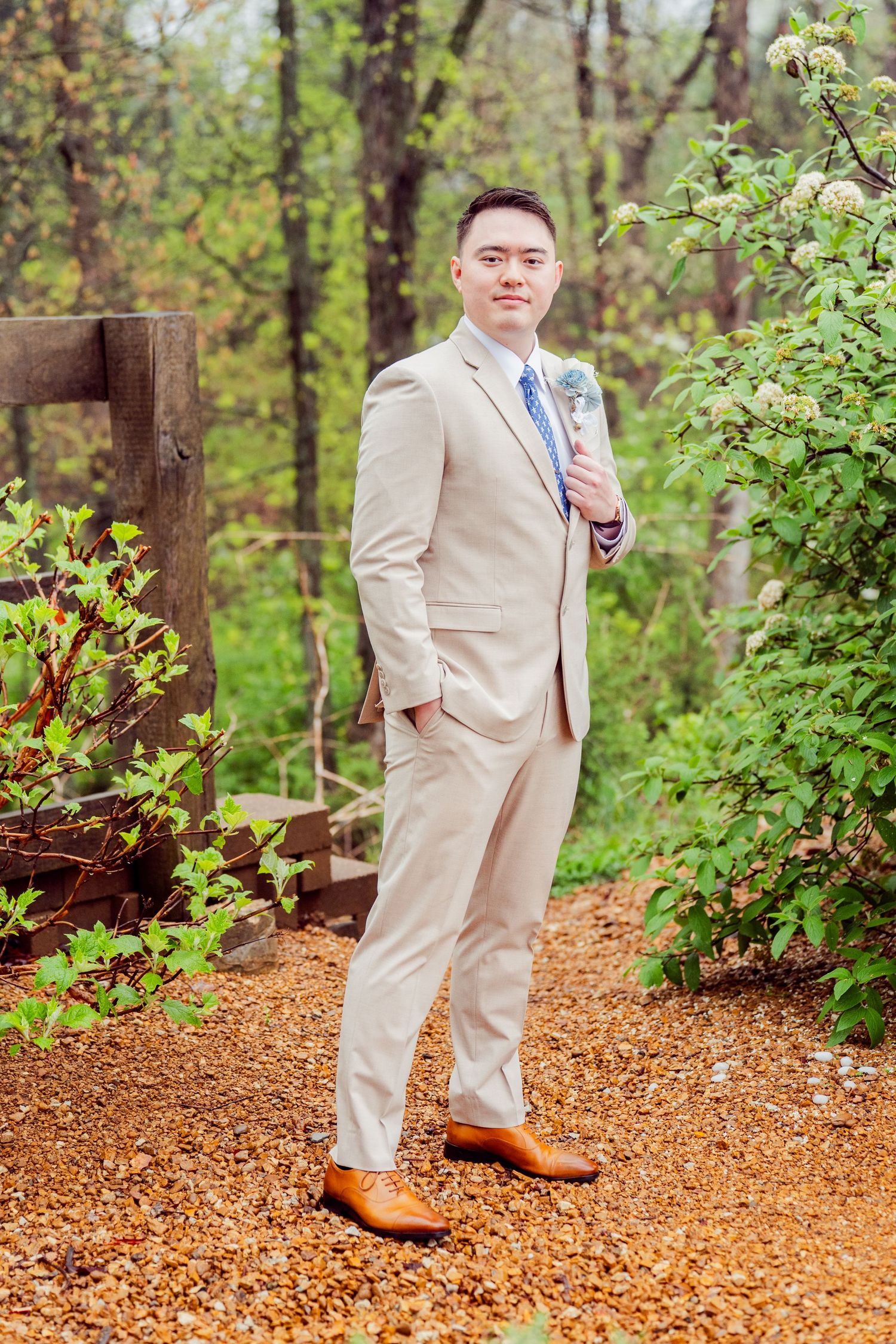 Groom in tan suit and blue tie poses on mulch path in wooded setting.
