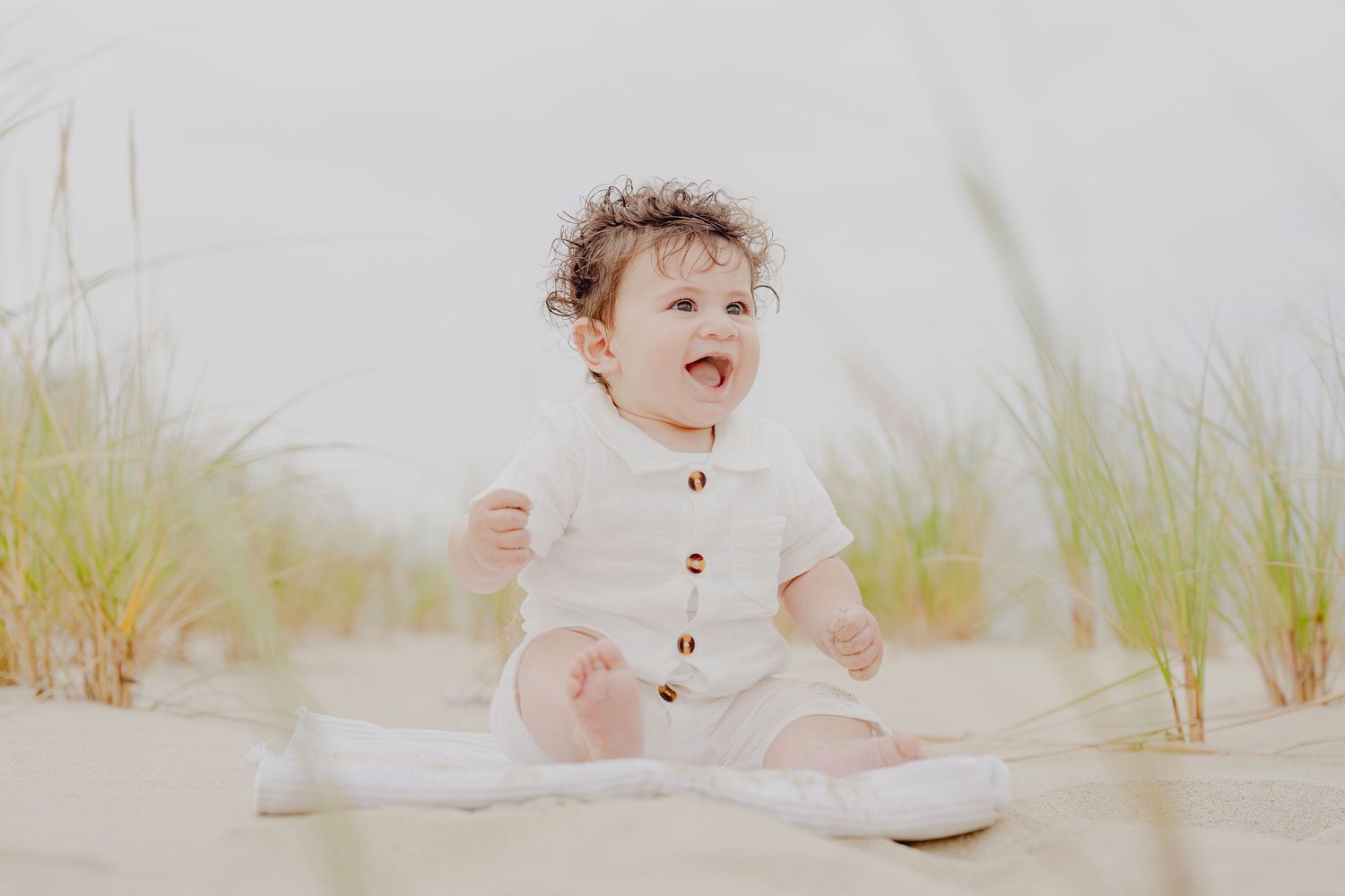 A happy baby in a white outfit sits on sandy beach dunes surrounded by grass, laughing joyfully.