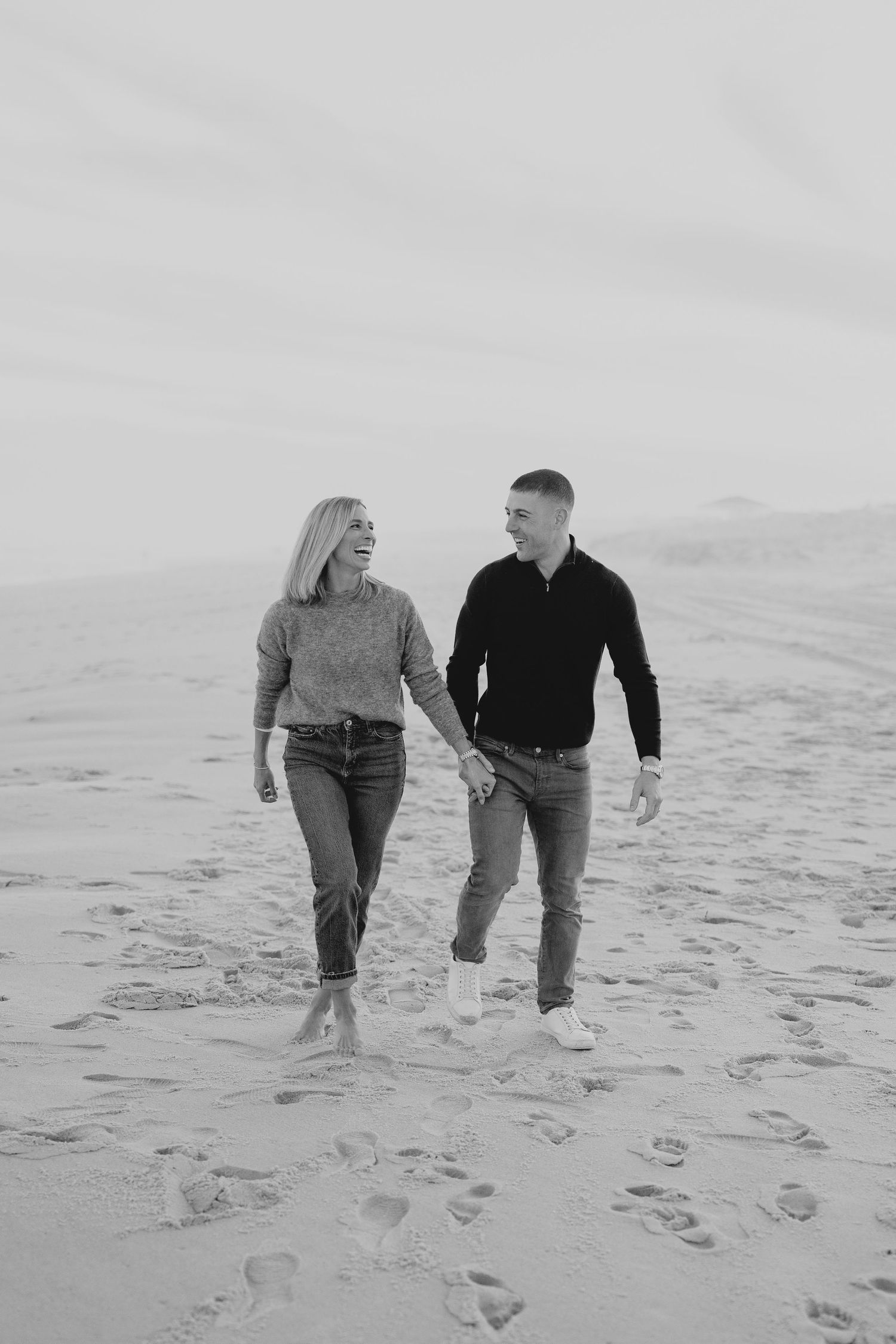 A couple walks together on a sandy beach leaving footprints in black and white photography.