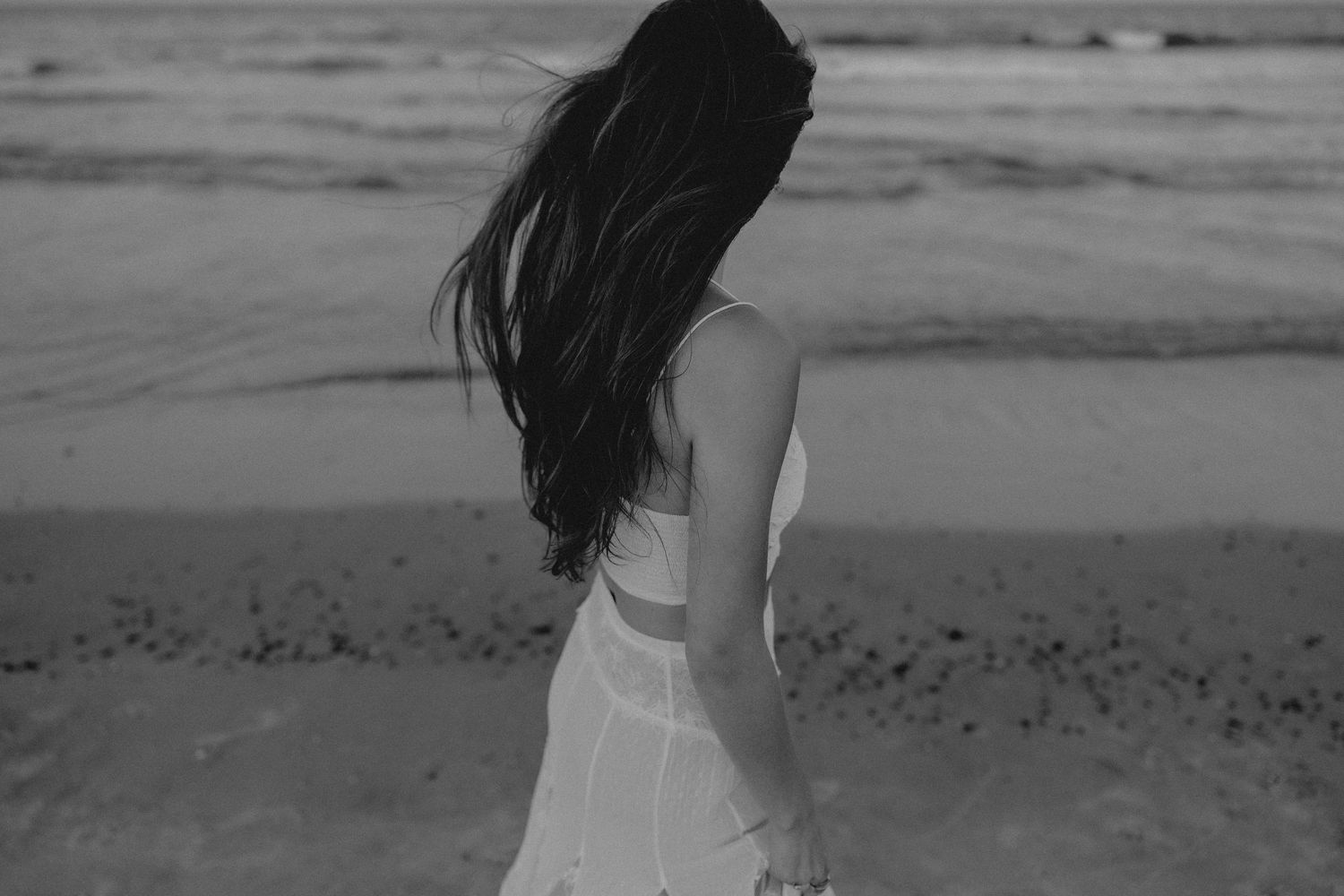 Black and white photo of a woman in a white dress standing on a beach with long dark hair blowing in the wind.