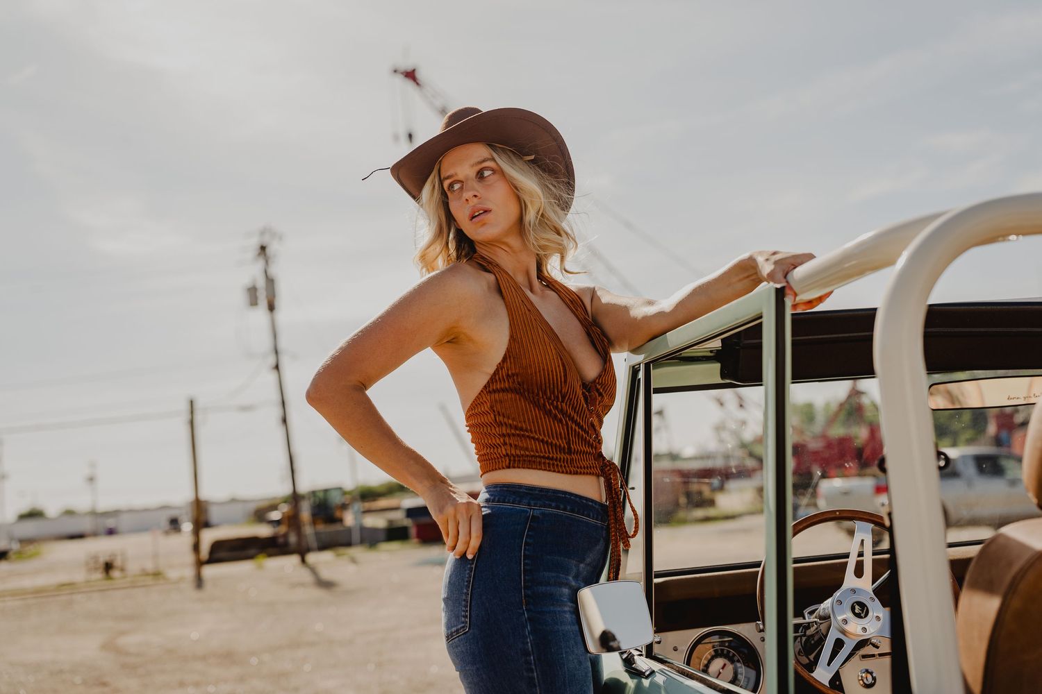 Blonde woman in rust halter top and jeans leaning on vintage mint Ford Bronco wearing a brown cowboy hat.