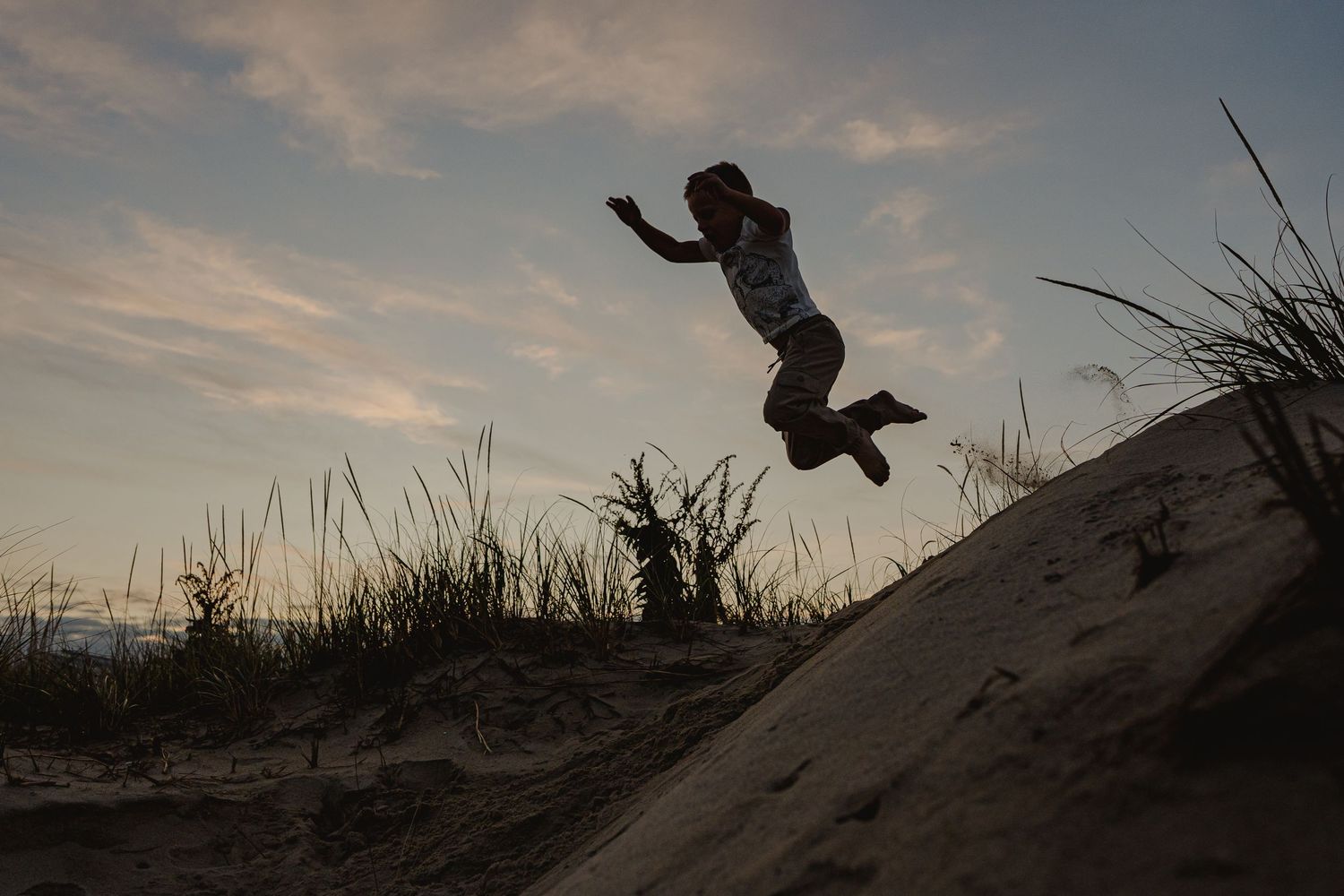 A child leaps joyfully off a sand dune at sunset, silhouetted against a dramatic sky near beach grass.