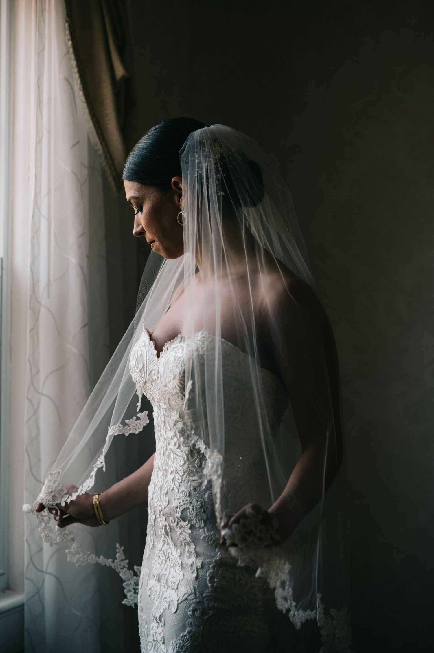 Moody window light illuminates a bride in lace dress and veil.