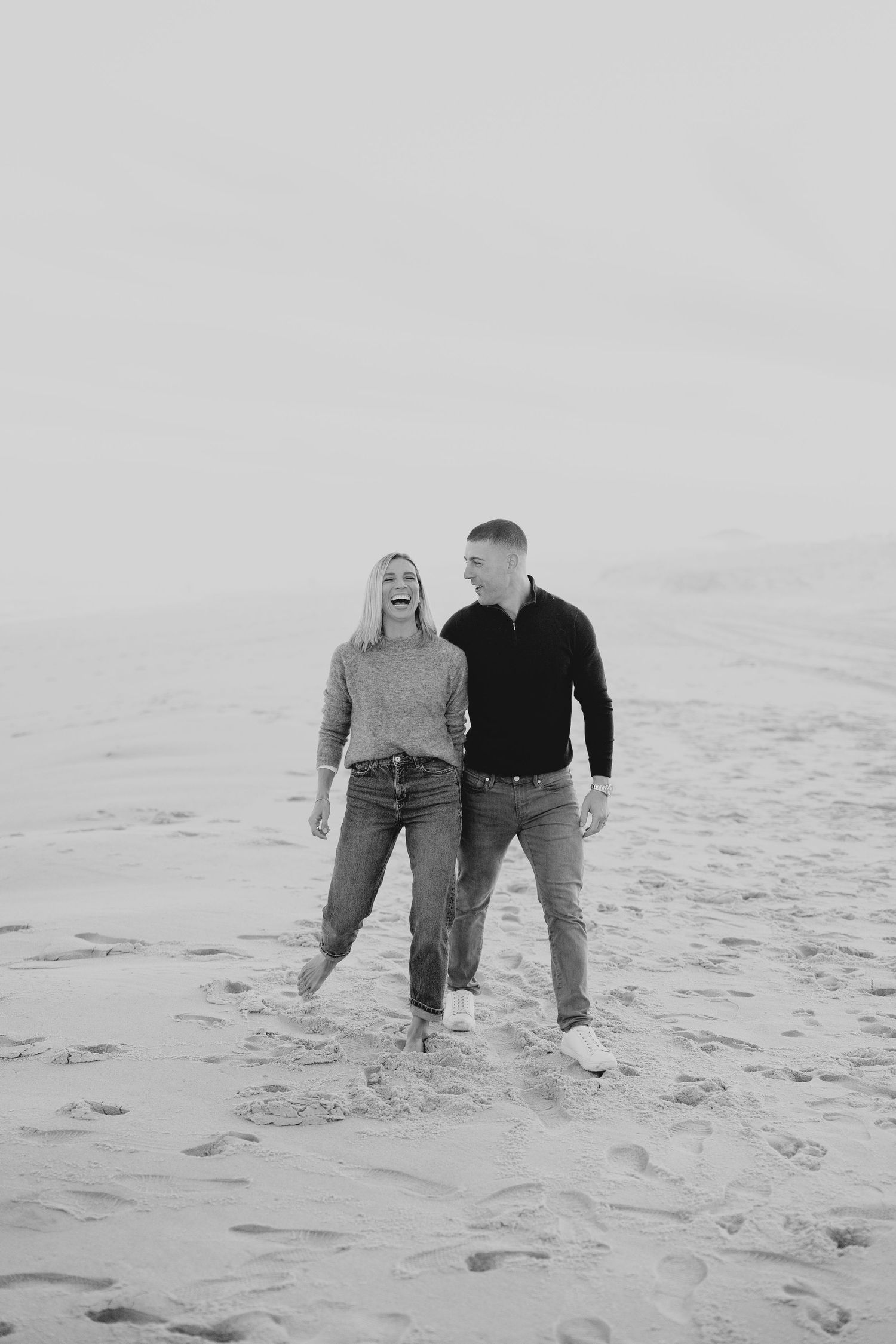 A couple walking together on a sandy beach in black and white, wearing casual clothing.