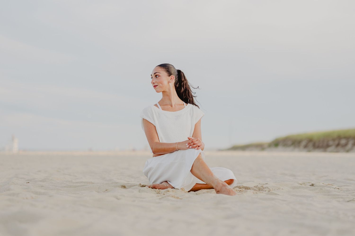 Woman in white dress sitting cross-legged on sandy beach, smiling peacefully with cloudy sky backdrop.