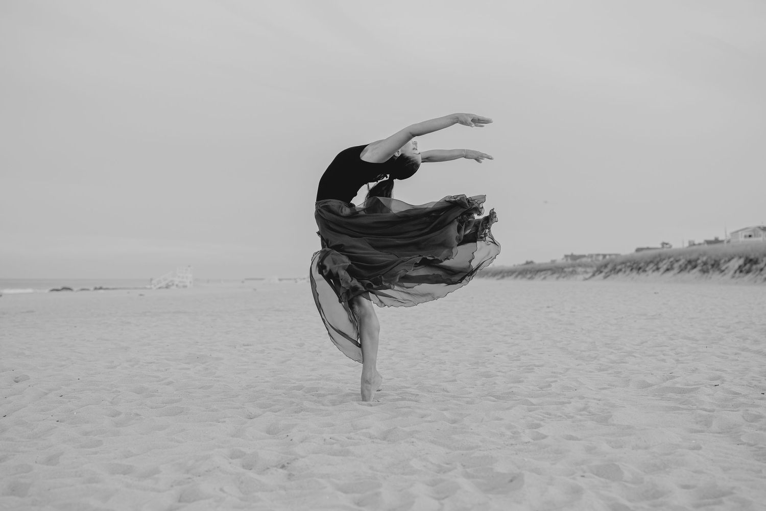 Black and white photo of a graceful dancer performing an arabesque on a sandy beach.