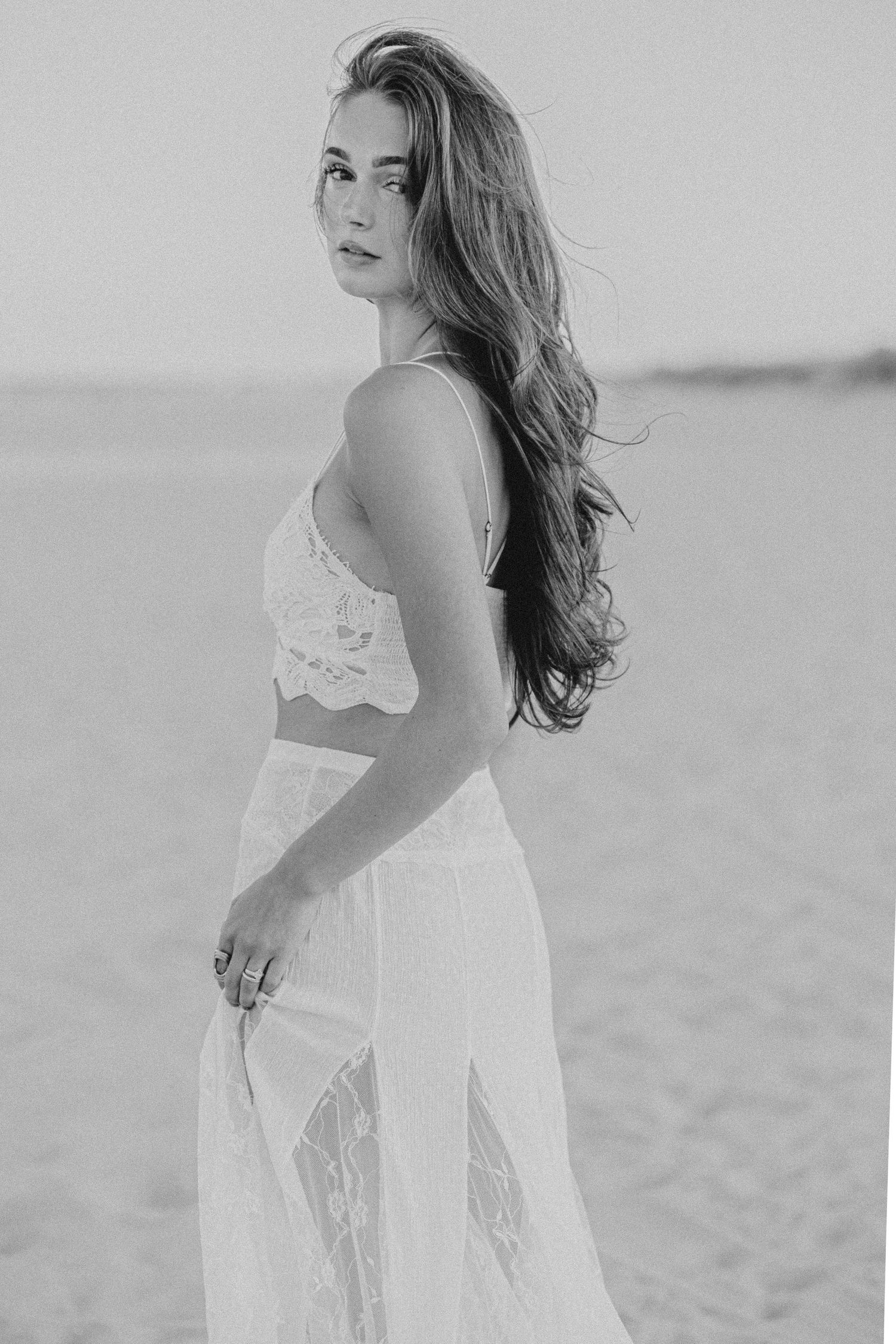 Woman in white lace crop top and skirt poses on sandy beach in black and white photo.