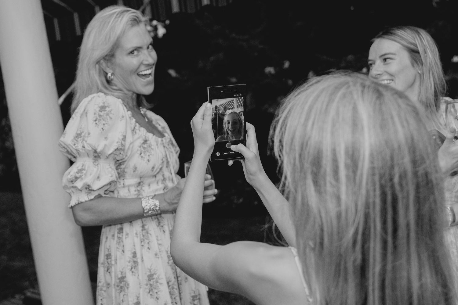 Woman in floral dress laughing as friend photographs her with smartphone at outdoor social gathering.