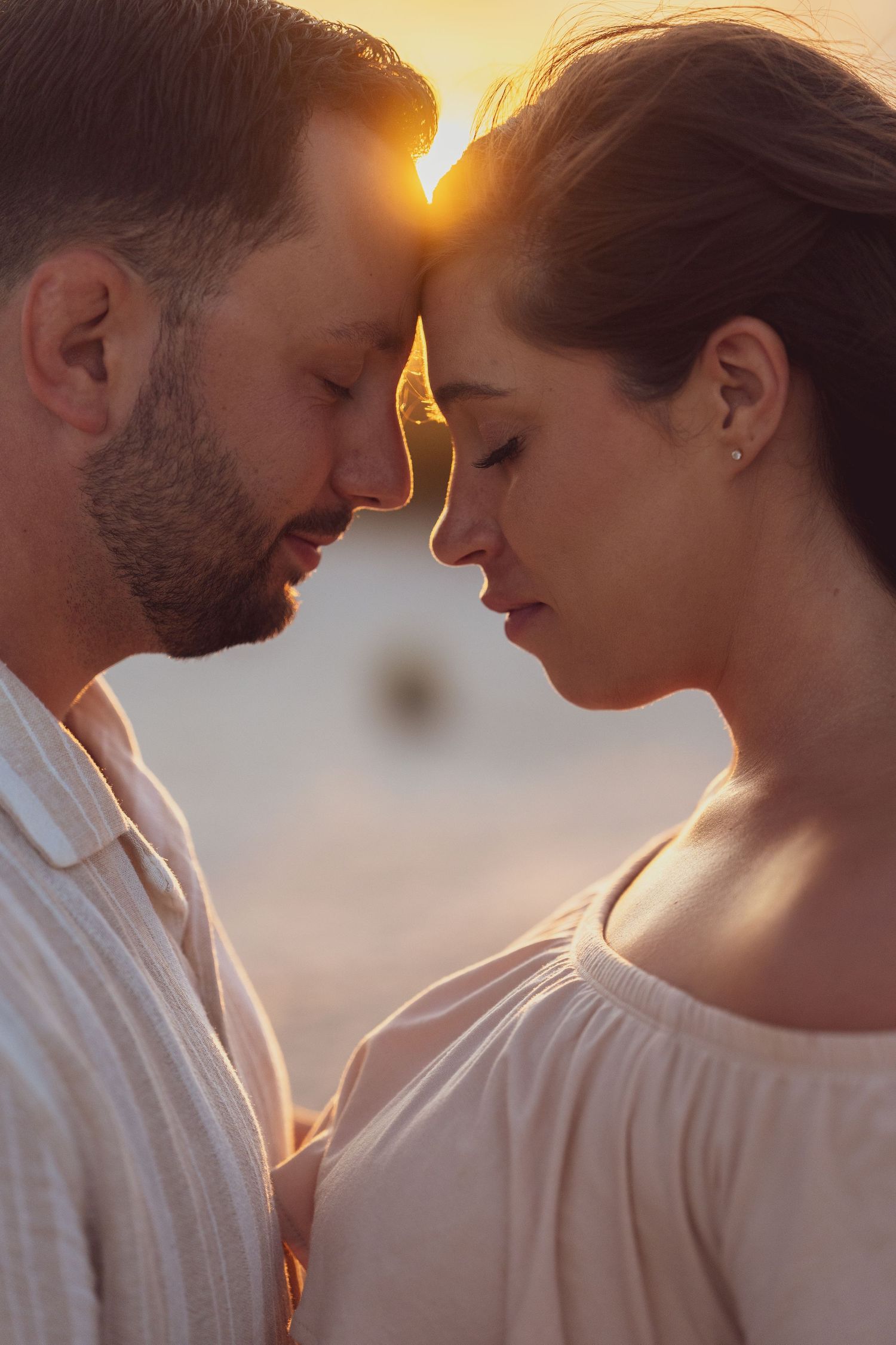 An intimate portrait of a couple in white clothing touching foreheads at golden hour.