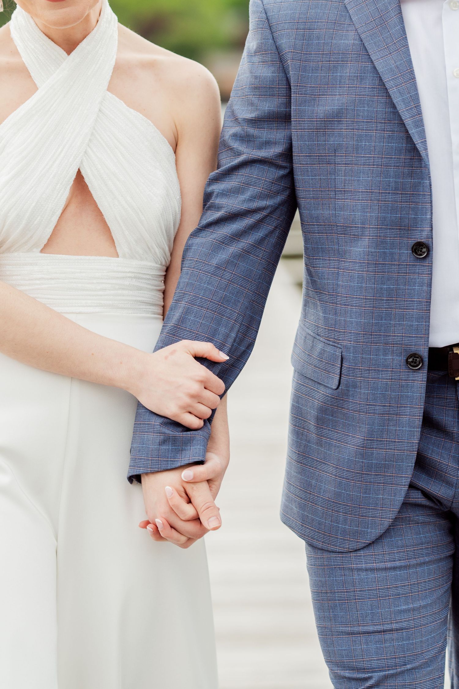 Close-up details of a bride in white dress and groom in blue suit holding hands.