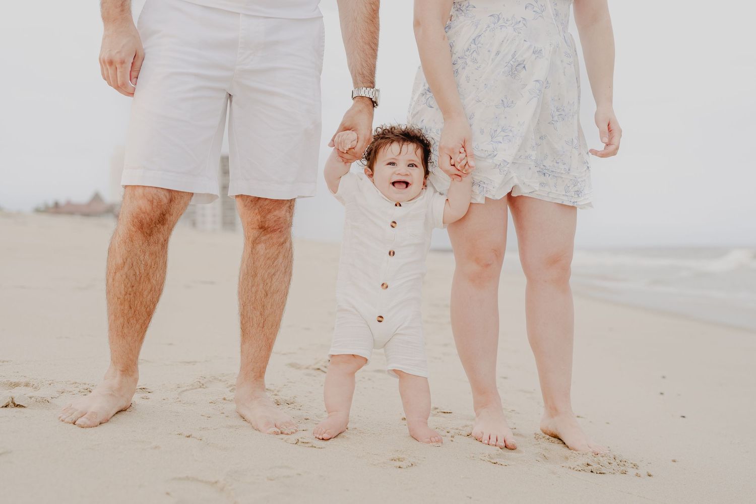 A happy baby takes first steps on a sandy beach, supported by parents in white outfits during a family photo session.