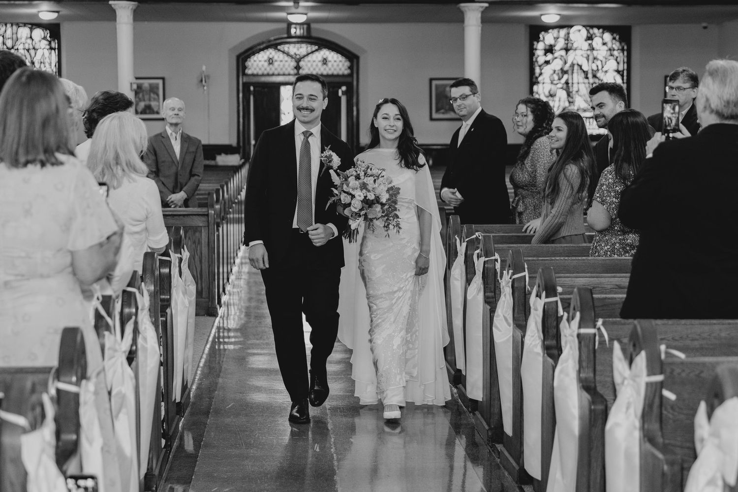Black and white photo of bride and groom walking down church aisle at wedding ceremony with guests watching.