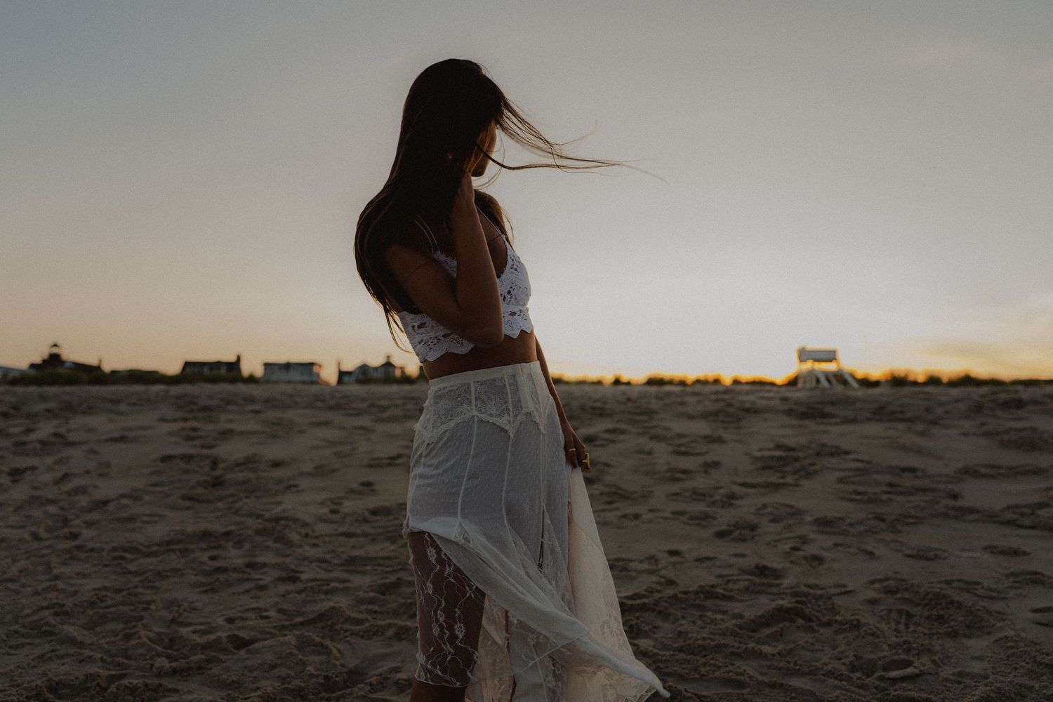 Silhouette of woman in white lace dress standing on beach at golden sunset.
