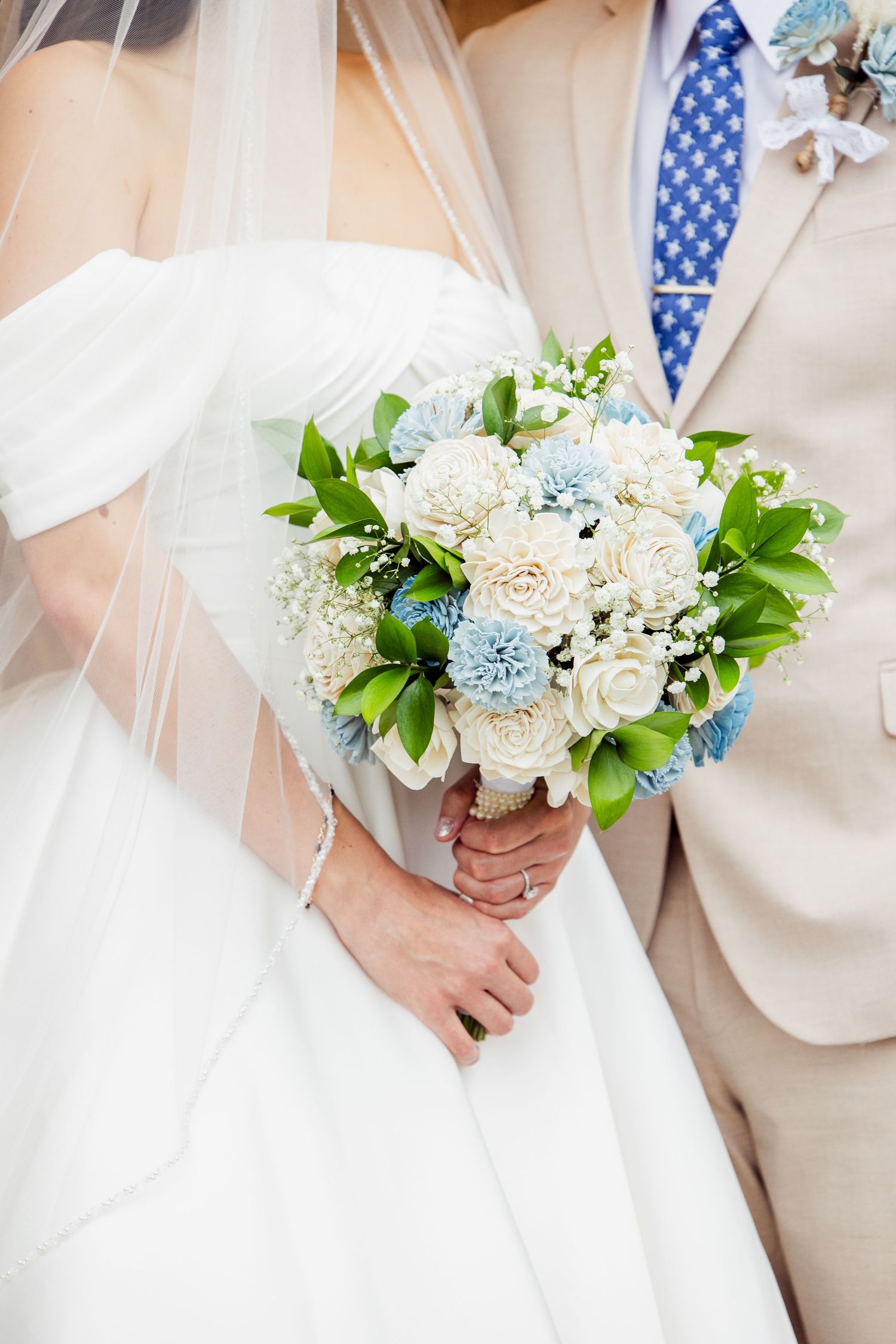 Wedding bouquet of white and blue flowers held against flowing white dress.