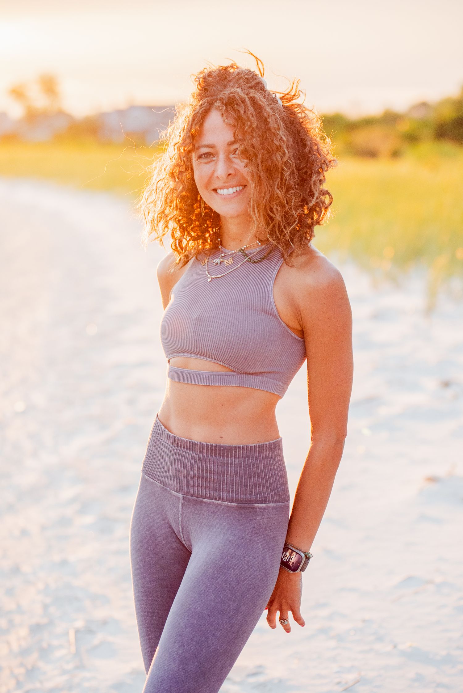 Athletic figure wearing purple workout attire poses on sandy beach at sunset.