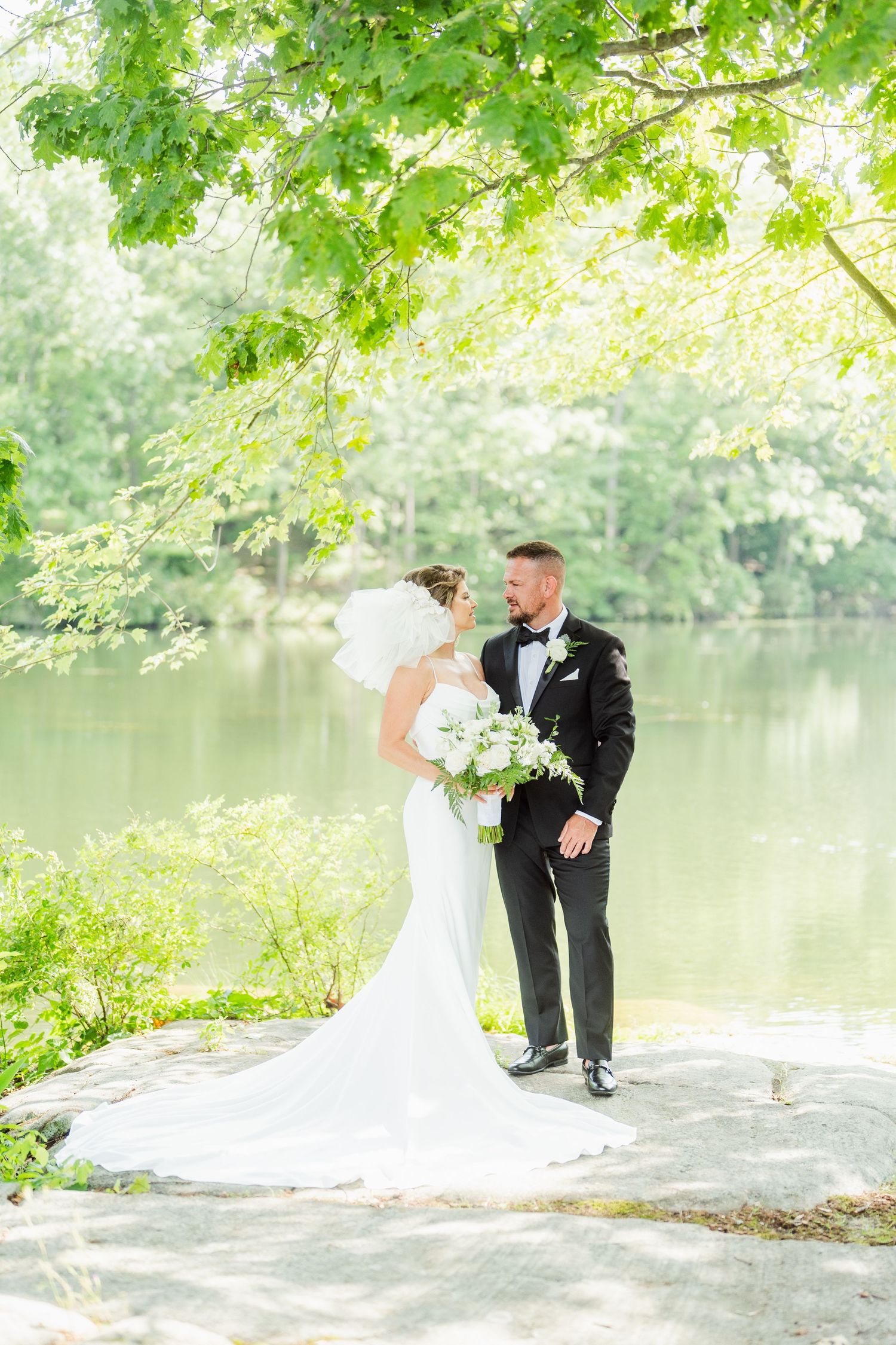 A romantic wedding photo series by a serene lake with lush green trees in the background.