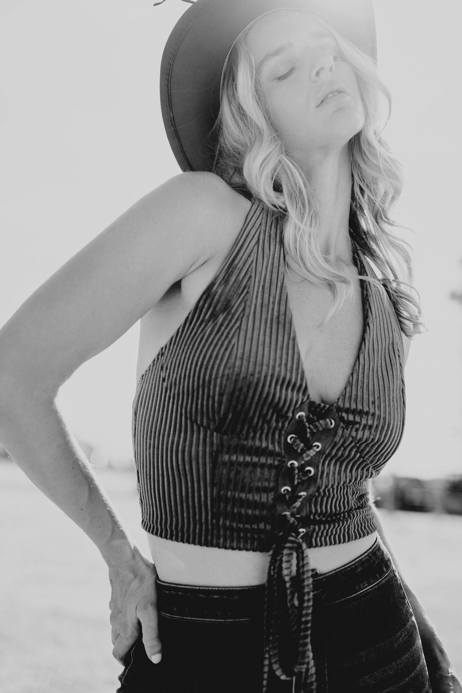 Black and white photo of a blonde woman in cowboy hat and lace-up crop top posing outdoors.