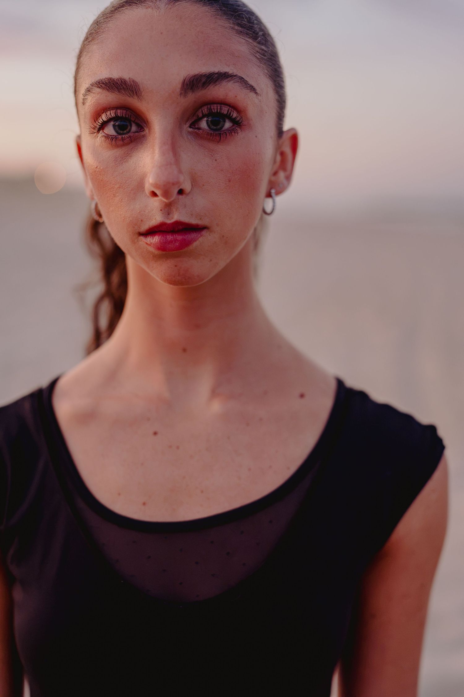 Young woman with freckles and red lips wearing a black mesh top poses outdoors at sunset.