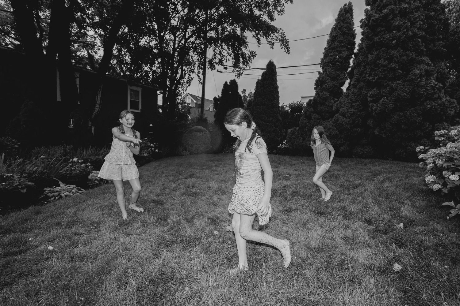 Black and white photo of three girls playing and running on a backyard lawn surrounded by trees and gardens.
