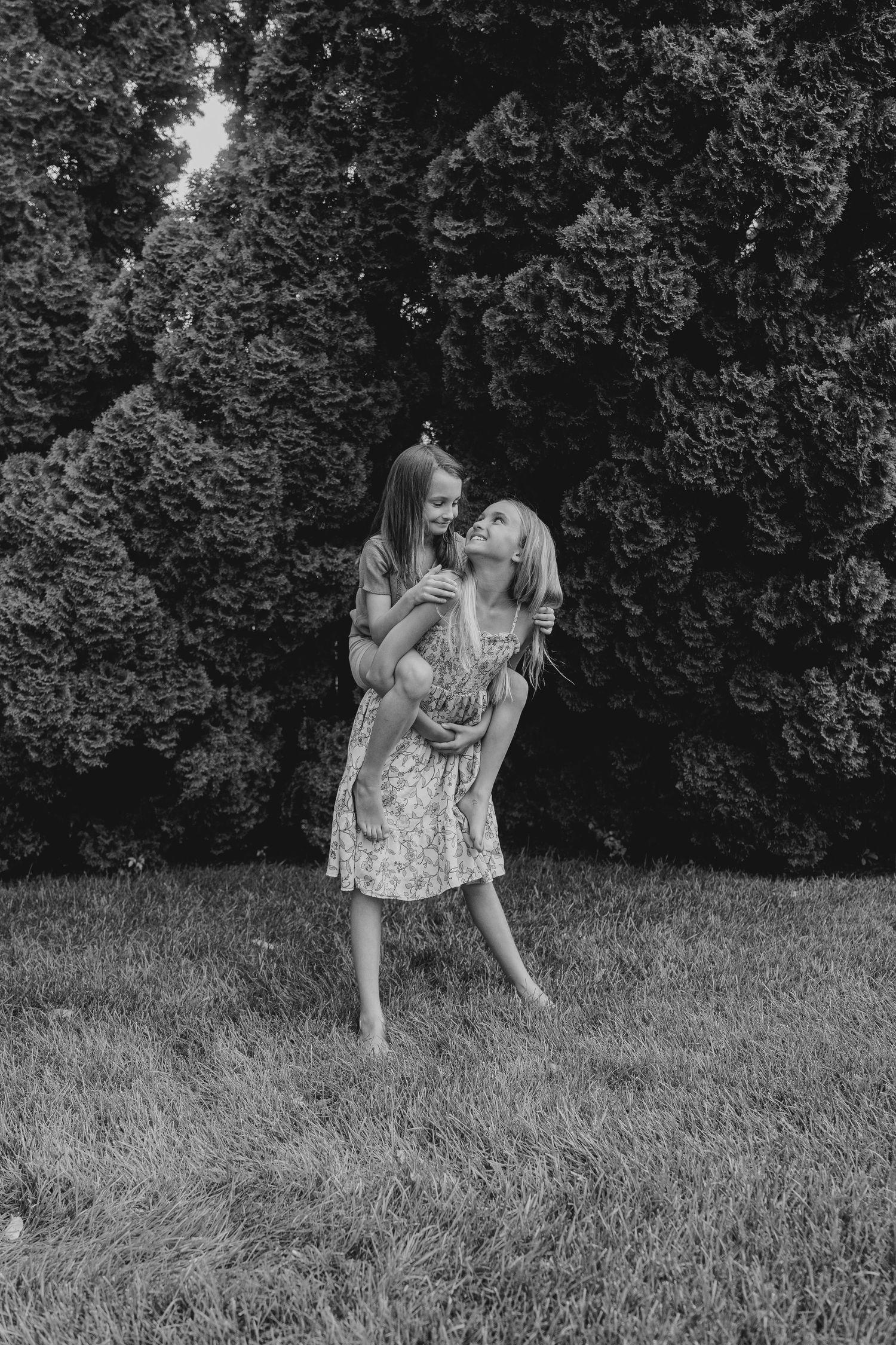 A woman and young girl laugh joyfully together outdoors, with the girl being held in front of lush evergreen trees.