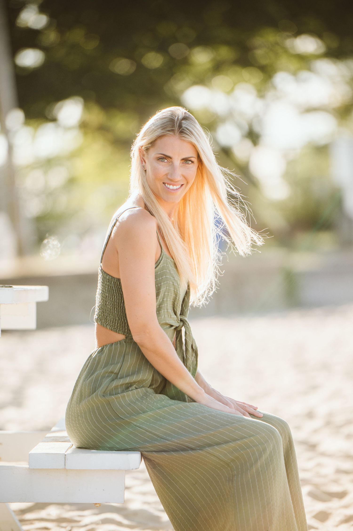 A woman in an olive green dress sits gracefully on a sandy beach during golden hour.