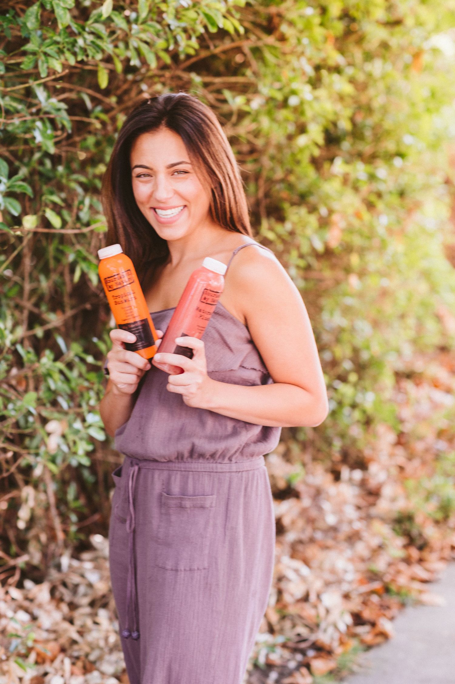 Woman in purple dress holds orange and pink cans while posing against green foliage background.