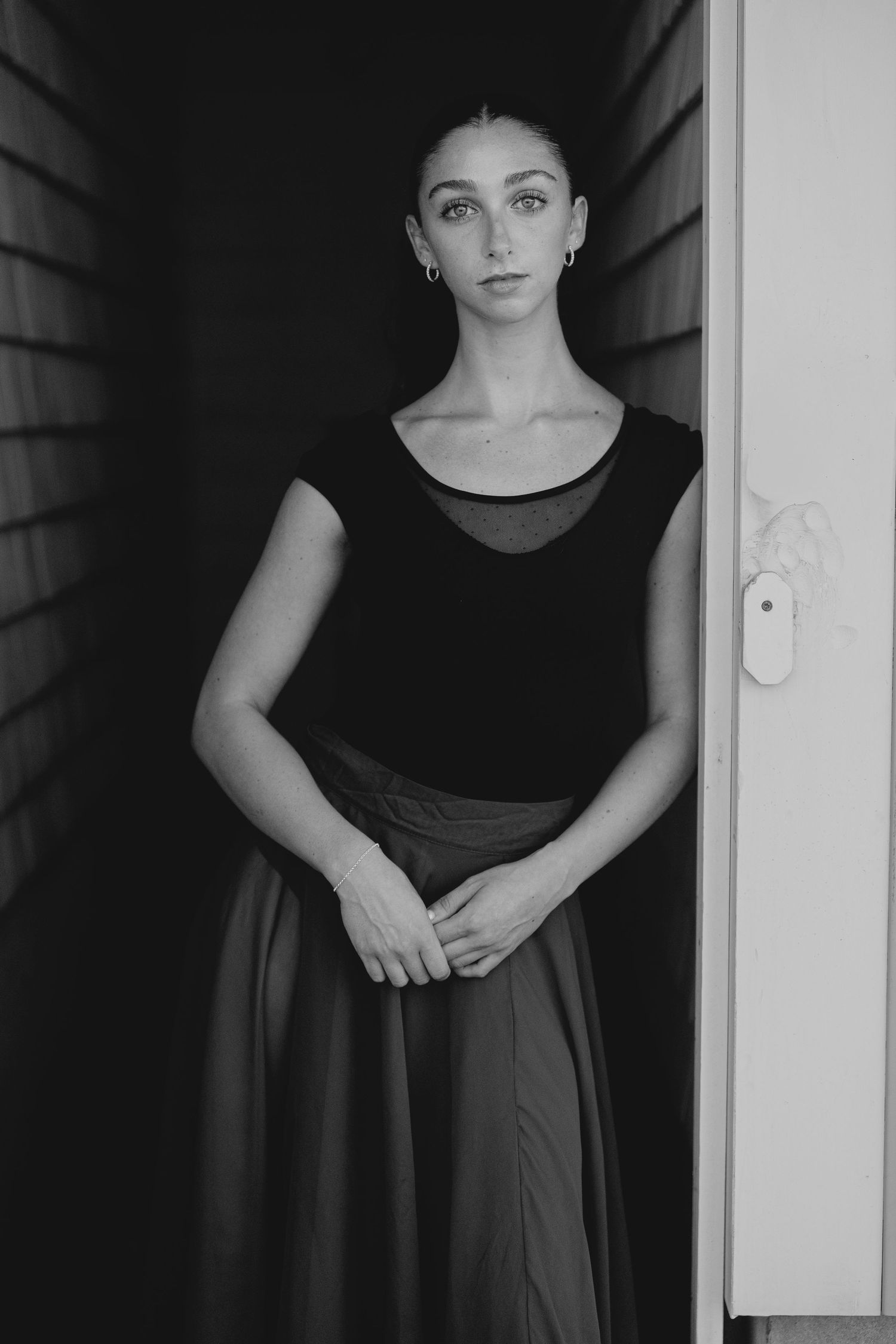 Black and white portrait of a young woman in a black top and skirt standing in a doorway.